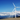 Eine Reihe von großen, weißen Windkraftanlagen (Windrädern) steht auf einem sandigen Küstenabschnitt vor dem Meer. Sie erstrecken sich in einer Linie in die Ferne, unter einem klaren, strahlend blauen Himmel mit weißen Kumuluswolken in der Ferne. Das Motiv symbolisiert erneuerbare Energien.