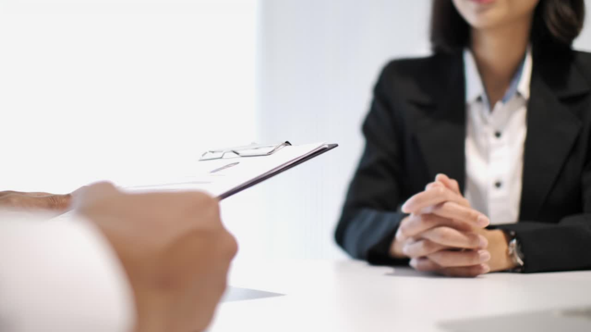Eine Szene, die ein Bewerbungsgespräch oder ein Geschäftstreffen darstellt. Im Vordergrund hält eine Person (nur die Hände sind sichtbar) ein Klemmbrett mit Dokumenten hin. Im Hintergrund sitzt eine unscharfe weibliche Person in formeller Kleidung (dunkler Anzug und weißes Hemd) am Tisch. Ihre Hände sind vor ihr gefaltet. Das Bild symbolisiert Personalwesen, Karriere oder berufliche Besprechungen.